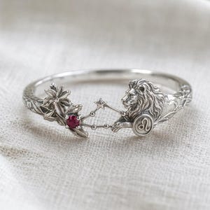 May include: A silver-toned bracelet featuring a detailed lion design, a red gemstone, and floral accents. The bracelet includes the Leo zodiac symbol and a constellation design. The bracelet is a piece of jewelry.