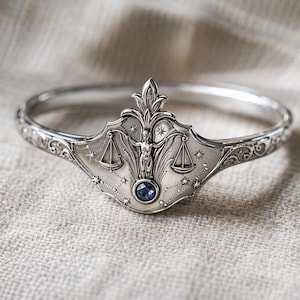 May include: A silver-toned bangle bracelet featuring a detailed Libra design. The centerpiece showcases a figure between scales, with a blue gemstone at the center. The bracelet has an ornate, celestial theme, with star and floral accents.