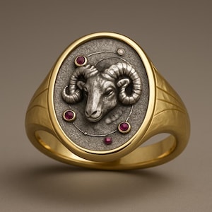 May include: A gold-toned ring with an oval face featuring a silver ram's head. The ring is accented with small red gemstones and a pearl-like detail. The band has a textured finish.