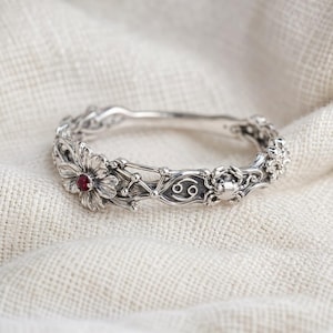 May include: A silver-toned bracelet with floral and celestial details. The bracelet features a flower with a red gemstone, a crab, and the Cancer zodiac symbol. The design is intricate and detailed, set against a neutral fabric background.