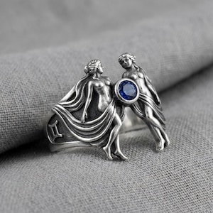 Könnte beinhalten: Ein silberfarbener Metallring mit zwei skulpturalen Figuren in fließenden Gewändern. Zwischen den Figuren befindet sich ein runder, tiefblauer Edelstein. An der Seite des Rings ist das Sternzeichen Zwillinge als Symbol eingraviert.
