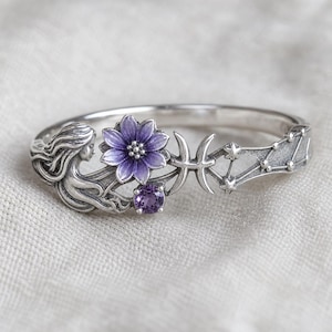Op de afbeelding: Een zilverkleurige armband met een zeemeermin, een paarse bloem, een Vissen-symbool en een sterrenbeeldontwerp. De bloem heeft een donkerpaars centrum en bloemblaadjes. Een kleine, ronde, paarse edelsteen is eronder geplaatst. De armband ligt op een witte ondergrond.