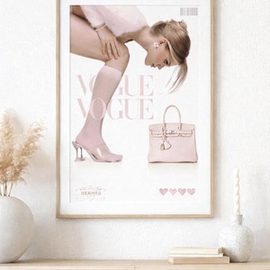 Könnte beinhalten: Gerahmter Druck mit einem rosa Vogue-Magazin-Cover-Design. Das Bild zeigt eine Frau, eine rosa Handtasche und den Text "Vogue". Der Druck wird an einer weißen Wand über einem Holzschrank angezeigt.