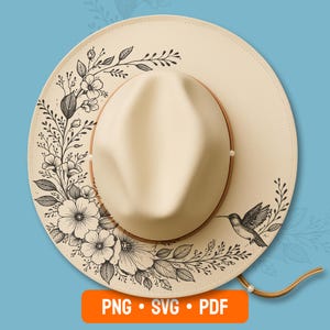 May include: Cream-colored fedora hat with a brown leather band and a chin strap. The hat's brim is decorated with a black floral and hummingbird design. The hat is a stylish accessory.