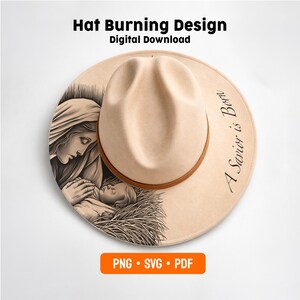 May include: A beige fedora hat featuring a black ink-style illustration of a woman holding an infant in a manger on the brim. The text A Savior is Born is printed on the right side. The hat has a brown band around the crown and a creased top.