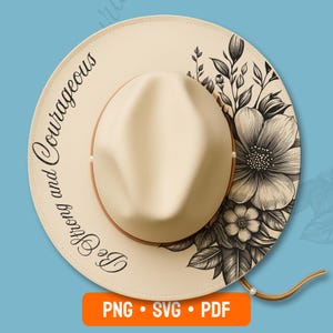 May include: A cream-colored fedora-style hat with a floral design on the brim. The hat features the text "Be Strong and Courageous" in a script font. A brown leather chin strap is attached. The hat is a stylish accessory.