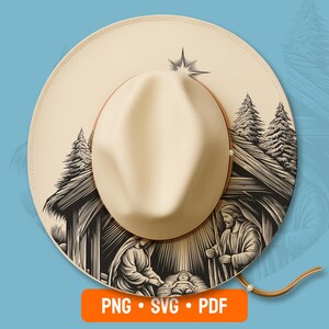 May include: A cream-colored wide-brim hat featuring a black and white line art illustration of a nativity scene with a star, pine trees, and a stable. The hat has a structured crown and a thin brown cord attached to the side of the brim.