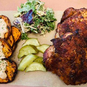 May include: A plate of food with two pieces of roasted chicken, sweet potato fries with a white sauce, a side salad with kale and a creamy dressing, and sliced pickles.
