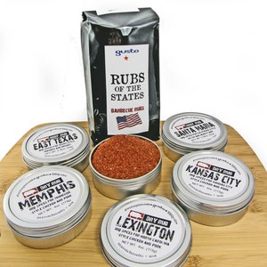 May include: A set of six metal tins containing different barbecue rubs. The tins are arranged on a wooden surface. The labels on the tins feature the names of different regions in the United States, such as Memphis, Kansas City, and Lexington. The labels also include the text "Dry Rub" and "BBQ Rubs".