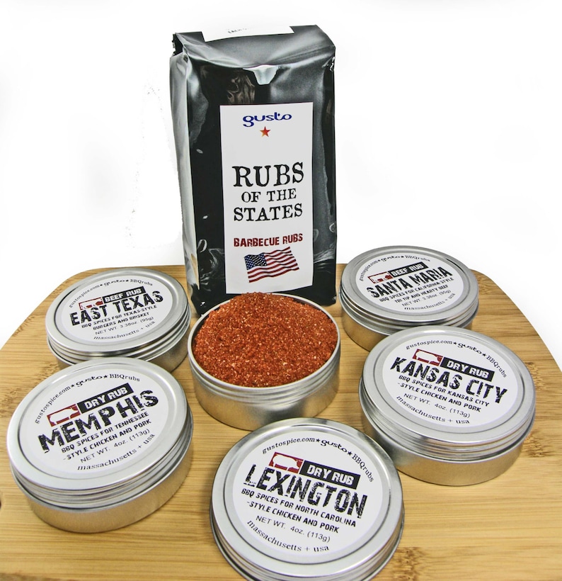 May include: A set of six metal tins containing different barbecue rubs. The tins are arranged on a wooden surface. The labels on the tins feature the names of different regions in the United States, such as Memphis, Kansas City, and Lexington. The labels also include the text "Dry Rub" and "BBQ Rubs".