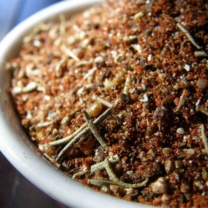 May include: A close-up of a white bowl filled with a mixture of brown, red, and white spices. The spices include dried herbs, peppercorns, and other seasonings.