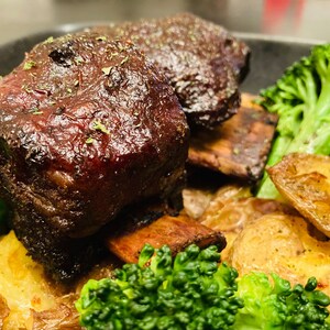 以下が含まれることがあります： 2つの大きな煮込みショートリブ、ローストポテト、ブロッコリーの花蕾が載った料理のプレートのクローズアップ。リブは濃いソースでコーティングされ、ポテトは黄金色です。