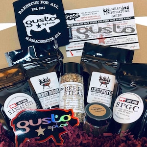 May include: A gift box filled with various spices and rubs for grilling and barbecuing. The box includes a black and white logo for "Gusto Spice" with the text "Barbecue for All" and "Massachusetts, USA". The spices are labeled with names like "Kansas City Dry Rub", "Hickory Burger", "Beef & Steak Rub Seasoning", "Lexington Style BBQ", and "SPGC Classic Beef and Pork Fit Rub".