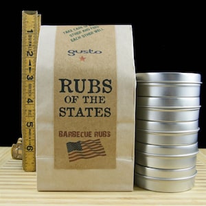 Puede incluir: Una bolsa de papel marr&oacute;n con una etiqueta que dice "Rubs of the States Barbecue Rubs". La etiqueta tambi&eacute;n presenta una peque&ntilde;a bandera estadounidense. Una pila de ocho latas de metal plateadas se encuentra frente a la bolsa.