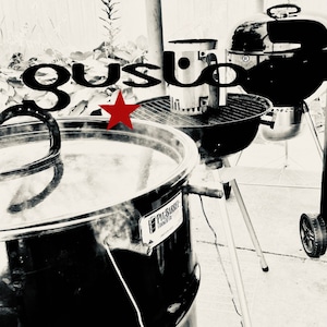 May include: A black metal smoker with a red star and the text "Pig Bar-B-Que Cooker Co." on the side. The smoker is on a tripod stand and there is a black grill in the background.
