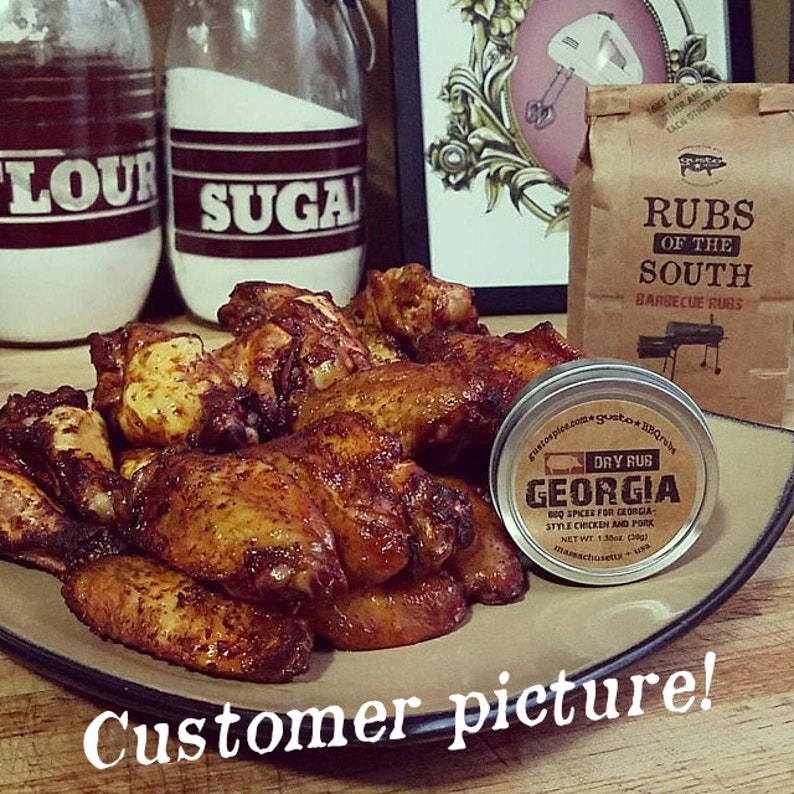 May include: Close-up of a plate of golden brown chicken wings, seasoned with a dry rub. The wings are piled high on a brown plate. A small tin of "Georgia Dry Rub" is visible in the foreground. The text "Customer picture!" is written in white on a black background at the bottom of the image.
