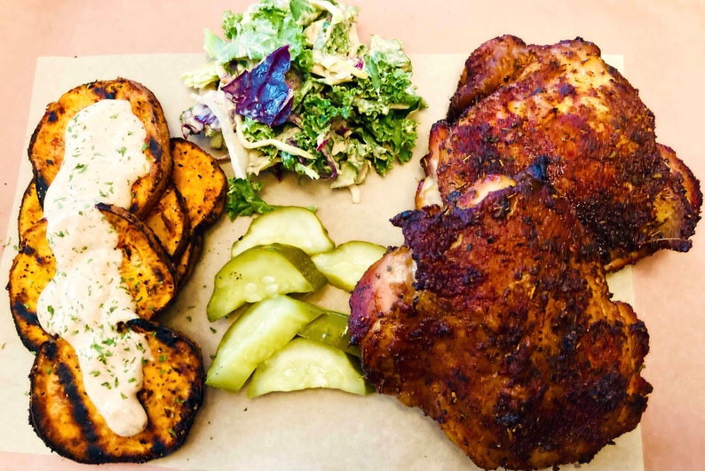 May include: A plate of food with two grilled chicken thighs, sweet potato fries with a white sauce, sliced pickles, and a side salad with kale and cabbage.