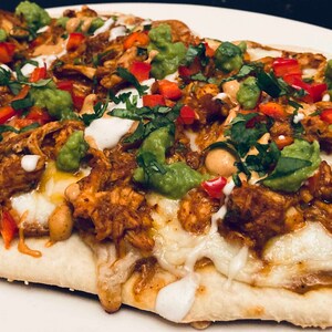 Puede incluir: Primer plano de una pizza de pan plano cubierta con pollo desmenuzado, guacamole, pimientos rojos, cilantro y una salsa blanca.