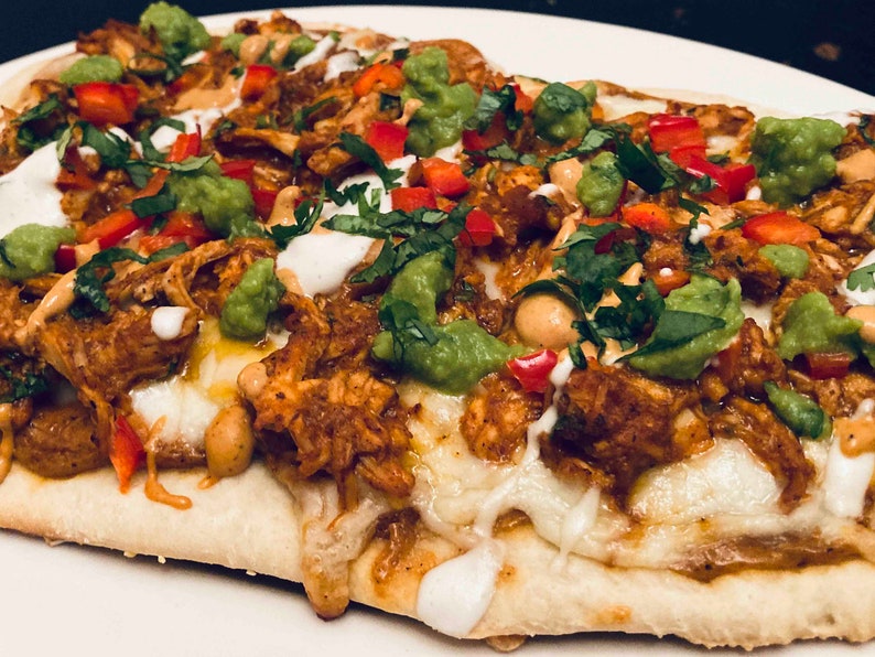 Puede incluir: Primer plano de una pizza de pan plano cubierta con pollo desmenuzado, guacamole, pimientos rojos, cilantro y una salsa blanca.