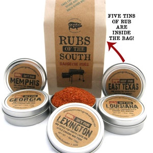 May include: Five metal tins of barbecue rubs, labeled with the names "Memphis", "Georgia", "East Texas", "Louisiana", and "Lexington". The tins are arranged in a circle around a brown paper bag with the text "Rubs of the South Barbecue Rubs".