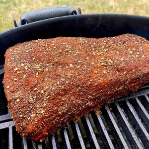 May include: A large piece of meat, likely brisket, is seasoned with a dry rub and placed on a black grill grate. The meat is covered in a thick layer of spices, including what appears to be garlic powder, paprika, and other herbs.