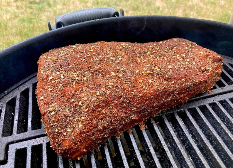 May include: A large piece of meat, likely brisket, is seasoned with a dry rub and placed on a black grill grate. The meat is covered in a thick layer of spices, including what appears to be garlic powder, paprika, and other herbs.