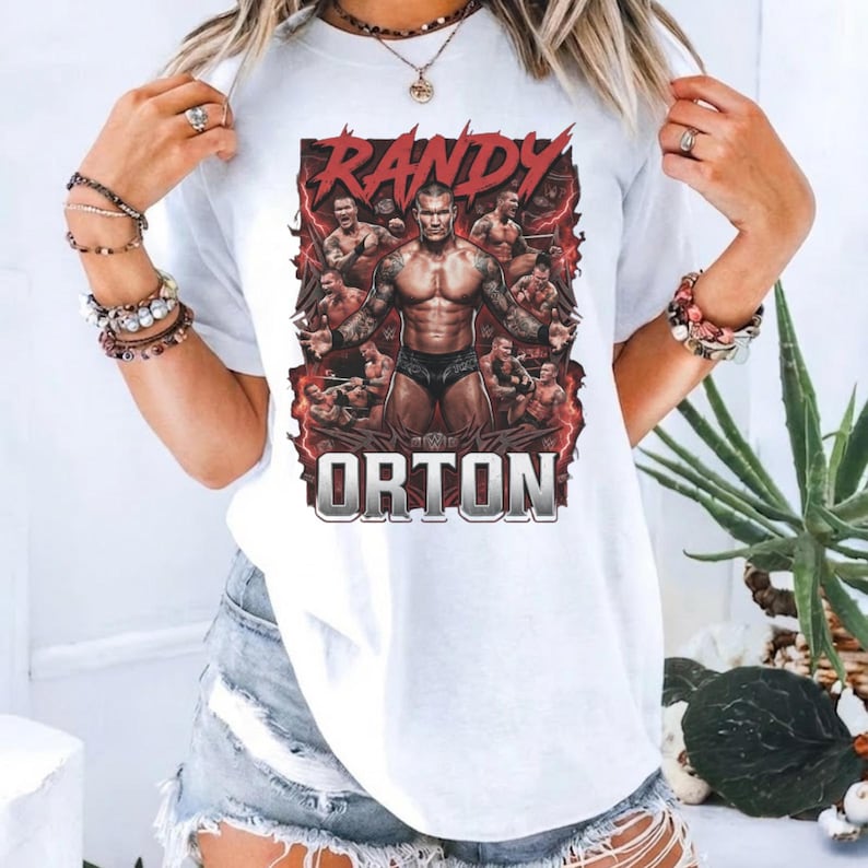 May include: A white t-shirt featuring a graphic print of professional wrestler Randy Orton in various poses. The design includes red lightning accents and the text RANDY ORTON in bold lettering. The shirt has a crew neckline and short sleeves.