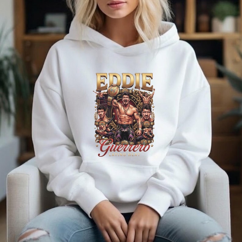 May include: A white hooded sweatshirt featuring a graphic print of professional wrestler Eddie Guerrero. The design includes the text EDDIE GUERRERO in gold and red, depicting the athlete holding a championship belt with multiple poses.