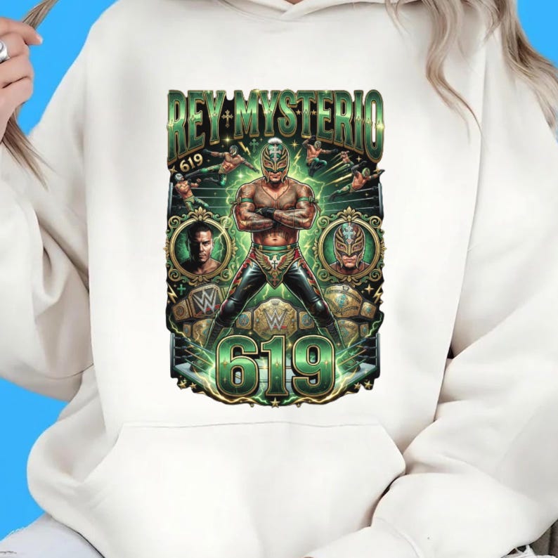 May include: A cream-colored hoodie featuring a graphic print of the wrestler Rey Mysterio in a mask and wrestling gear. The design includes the text "REY MYSTERIO" and "619" in green, along with smaller images of the wrestler and championship belts.