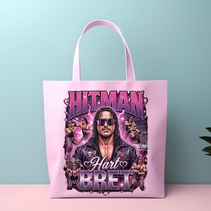 May include: A light pink tote bag featuring a graphic print of professional wrestler Bret Hart. The design includes the text HITMAN and BRET HART, multiple images of the wrestler in action, and purple lightning accents against a pink background.