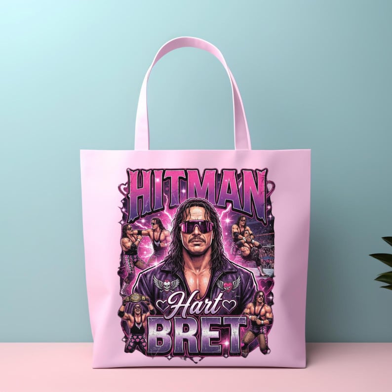 May include: A light pink tote bag featuring a graphic print of professional wrestler Bret Hart. The design includes the text HITMAN and BRET HART, multiple images of the wrestler in action, and purple lightning accents against a pink background.