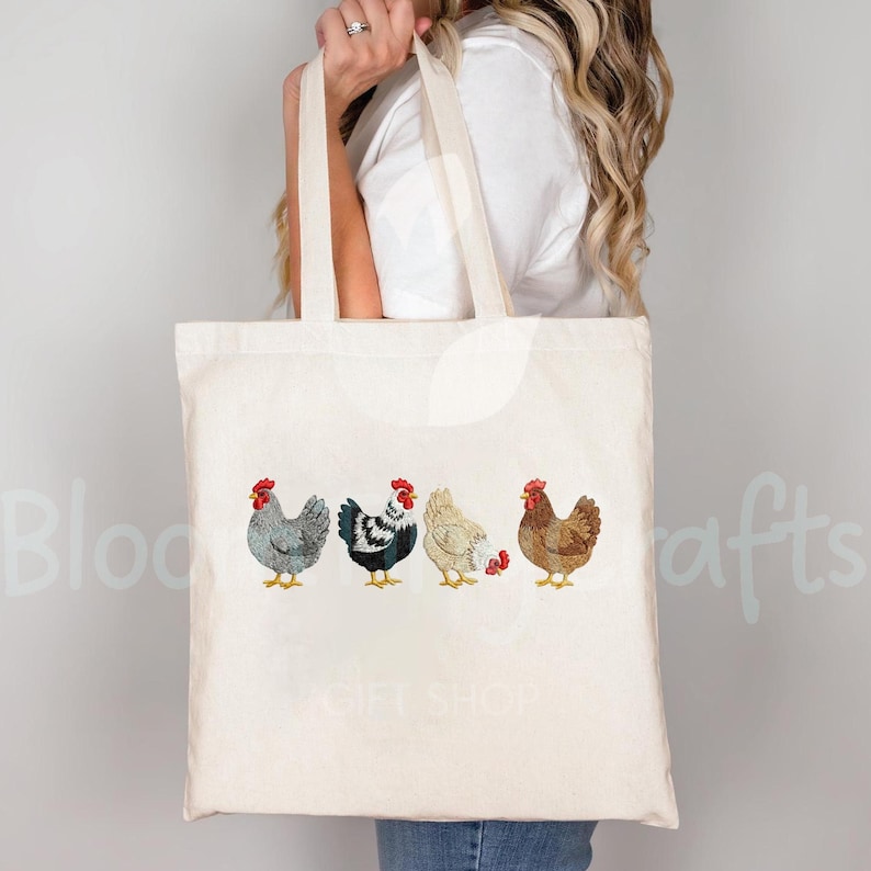 May include: A cream-colored tote bag featuring four embroidered chickens in a row. The chickens are in shades of gray, black and white, cream, and brown, each with a red comb. The bag is held by a person.