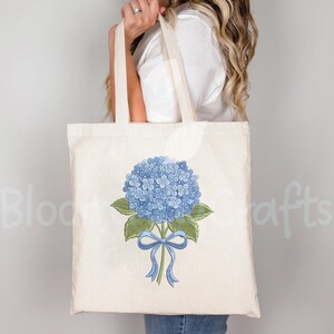 May include: A cream-colored tote bag featuring a blue hydrangea flower with green leaves and a blue ribbon. The bag has long handles and is held by a person. The words "Bloom Crafts" are partially visible on the left side of the bag.