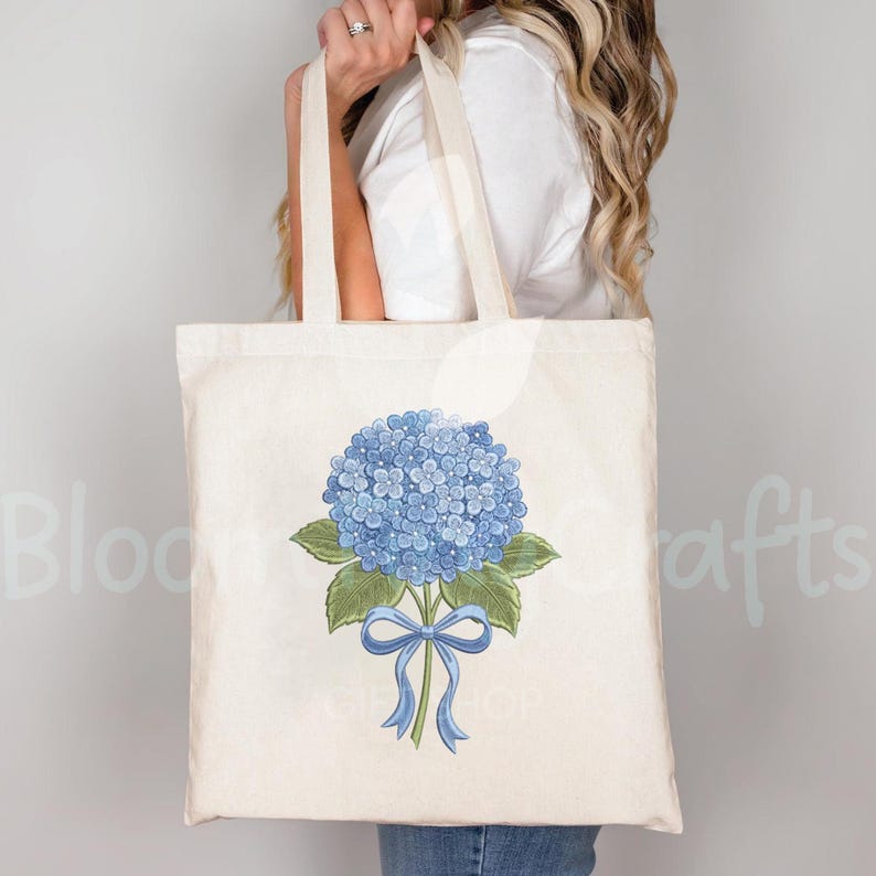 May include: A cream-colored tote bag featuring a blue hydrangea flower with green leaves and a blue ribbon. The bag has long handles and is held by a person. The words "Bloom Crafts" are partially visible on the left side of the bag.