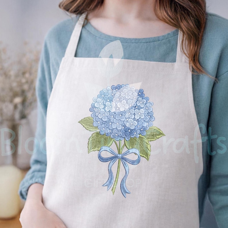 May include: A white apron featuring a blue hydrangea flower illustration with green leaves and a blue ribbon. The apron is made of a light-colored fabric and has a simple design, suitable for kitchen or craft use.