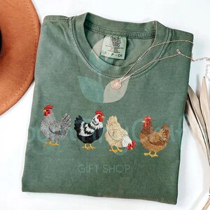 May include: An olive green t-shirt with embroidered chickens in a row. The chickens are in various colors, including gray, black and white, tan, and brown, each with a red comb. A silver necklace with a pendant is on the shirt.