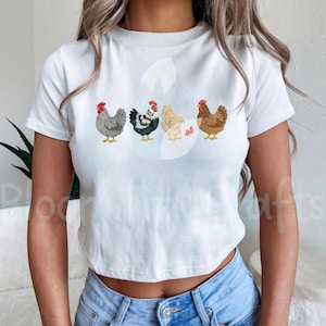 May include: White t-shirt featuring an embroidered design of four chickens in a row. The chickens are in various colors, including gray, black and white, cream, and brown, each with a red comb. The shirt is a cropped style.