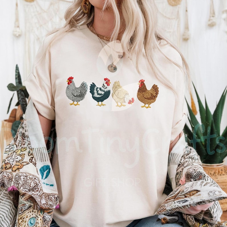 May include: Cream-colored t-shirt featuring an embroidered design of five chickens in various colors. The chickens have red combs and yellow feet. The shirt has a relaxed fit and short sleeves.