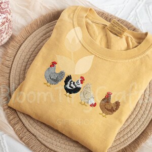 May include: Mustard yellow sweatshirt featuring embroidered chickens in various colors. The chickens have red combs and are arranged in a row across the front of the shirt. The text "Bloom Tree Craft" and "Gift Shop" are visible.