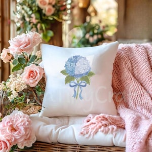 May include: A white square throw pillow featuring a blue and white hydrangea floral design with green leaves and a blue ribbon. The pillow is placed on a white cushion, with pink roses and a pink blanket in the background.