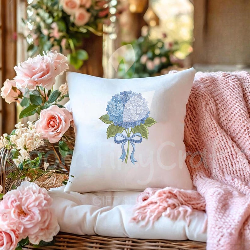 May include: A white square throw pillow featuring a blue and white hydrangea floral design with green leaves and a blue ribbon. The pillow is placed on a white cushion, with pink roses and a pink blanket in the background.