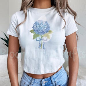 May include: White t-shirt featuring a watercolor illustration of a blue hydrangea flower bouquet tied with a blue ribbon. The floral design includes green leaves and a light blue bow. The shirt is a crew neck style.