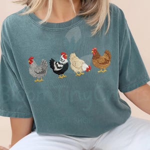 May include: Teal-colored t-shirt featuring an embroidered design of four chickens in a row. The chickens are in various colors, including gray, black and white, tan, and brown, with red combs and yellow feet. The shirt is a casual style.