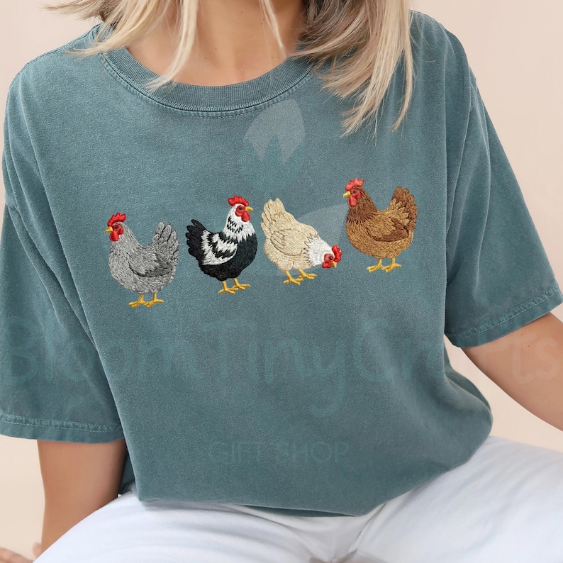 May include: Teal-colored t-shirt featuring an embroidered design of four chickens in a row. The chickens are in various colors, including gray, black and white, tan, and brown, with red combs and yellow feet. The shirt is a casual style.