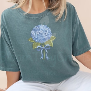 May include: Teal-colored t-shirt featuring a detailed illustration of a blue hydrangea flower with green leaves and a blue ribbon. The floral design is centered on the shirt, creating a classic and elegant look. The shirt is made of a soft material.