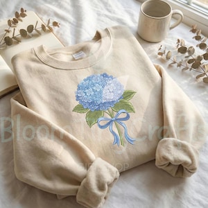May include: A beige crewneck sweatshirt featuring a blue hydrangea bouquet design. The floral illustration includes a cluster of blue flowers, green leaves, and a blue ribbon. The sweatshirt has a relaxed fit and long sleeves.