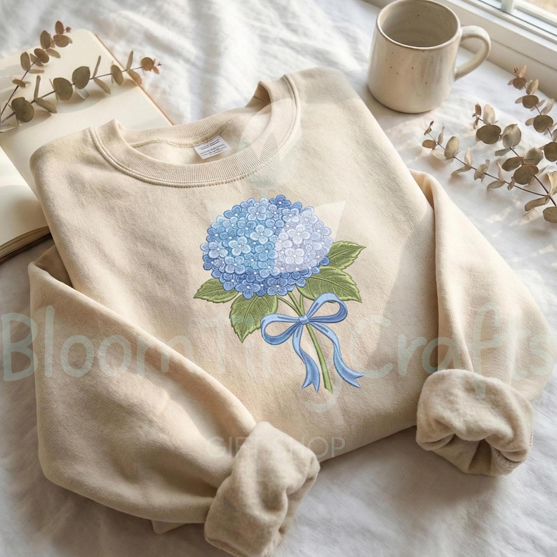 May include: A beige crewneck sweatshirt featuring a blue hydrangea bouquet design. The floral illustration includes a cluster of blue flowers, green leaves, and a blue ribbon. The sweatshirt has a relaxed fit and long sleeves.