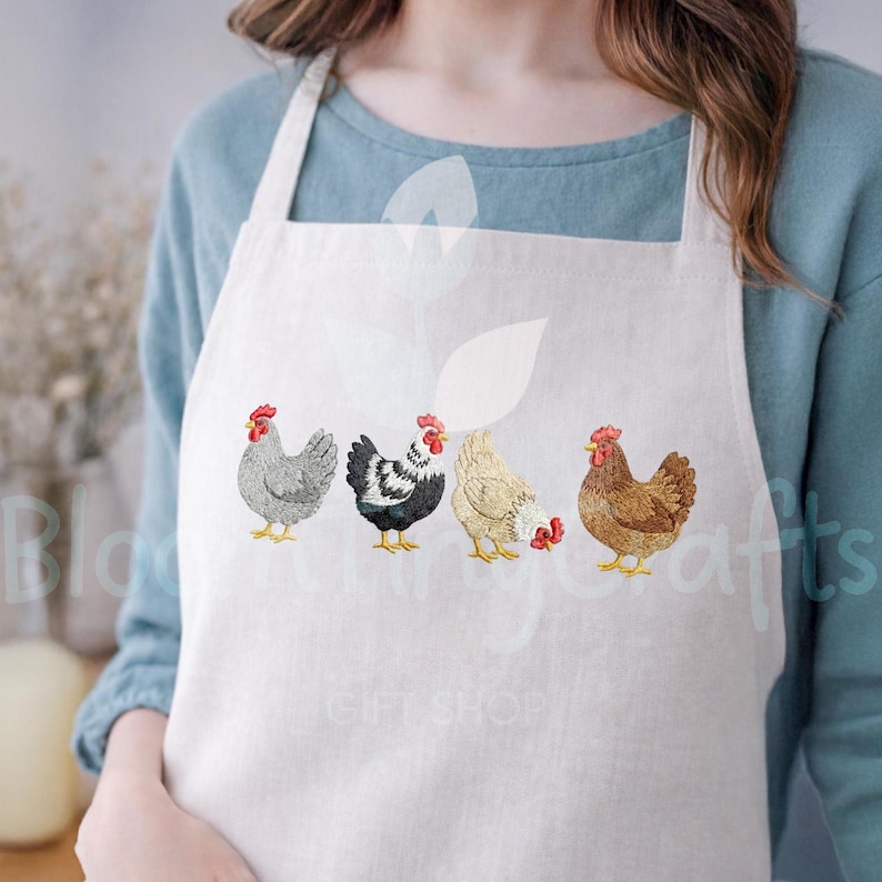 May include: A white apron featuring embroidered chickens in various colors. The chickens have red combs and wattles, with detailed feathering. The apron is worn by a person, with the text "GIFT SHOP" visible.