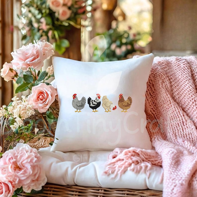 May include: White square pillow featuring embroidered chickens in various colors. The chickens are gray, black and white, light yellow, and brown, each with a red comb. The pillow is on a wicker chair with pink roses and a pink blanket.