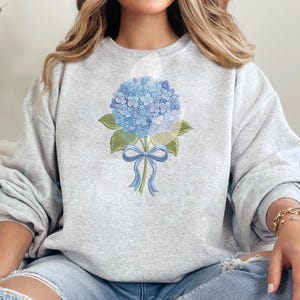 May include: A light gray sweatshirt featuring a blue hydrangea bouquet design. The floral illustration includes a cluster of blue flowers, green leaves, and a blue ribbon. The sweatshirt is a comfortable, casual style.